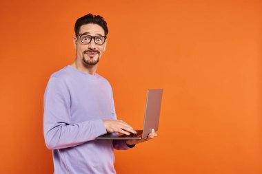 Gözlüklü ve mor kazaklı adam turuncu arka planda dizüstü bilgisayar kullanıyor.