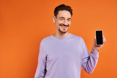 Gözlüklü ve mor kazaklı mutlu bir adam elinde turuncu arka planda boş ekranlı bir akıllı telefon tutuyor.