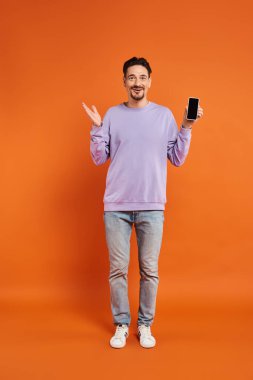 amazed man in glasses and purple sweater holding smartphone with blank screen on orange background clipart