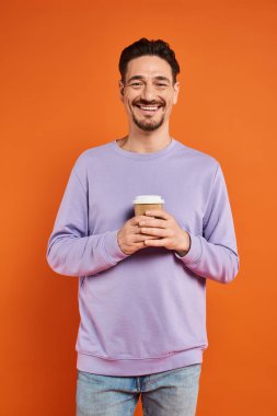 Neşeli ve sakallı bir adam elinde kahve bardağıyla turuncu arka planda gezmek için kağıt bardak tutuyor.