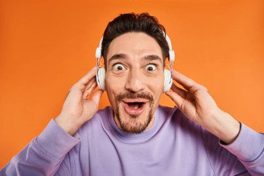 Kablosuz kulaklıklı sakallı adam müzik dinliyor ve turuncu arka plandaki kameraya bakıyor.