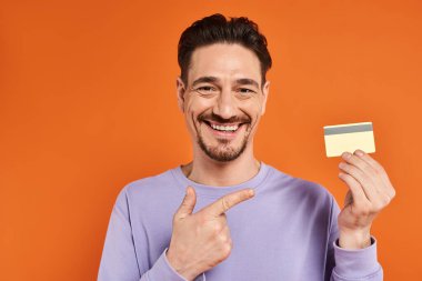 cheerful man with beard smiling and pointing with finger at credit card on orange background clipart