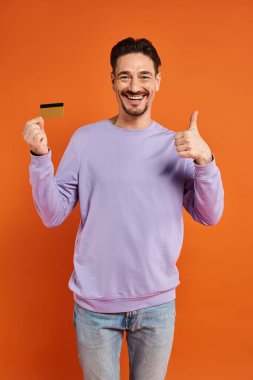 cheerful man with beard shoeing thumb up and holding credit card on orange background, like clipart
