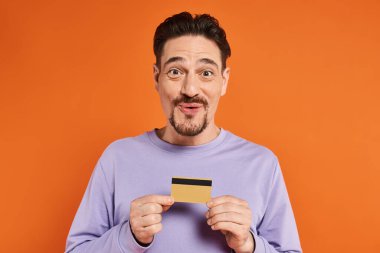 cheerful man with beard smiling and holding credit card on orange background, looking at camera clipart