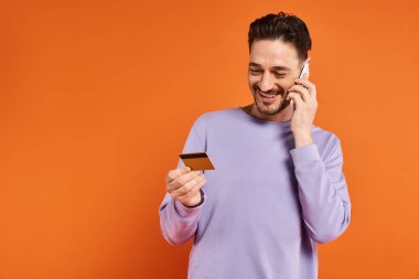 cheerful man smiling and holding credit card while talking on smartphone on orange background clipart