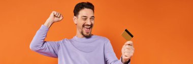 excited man with beard smiling and holding credit card on orange background, rejoicing banner clipart