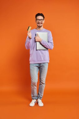 Gözlüklü mutlu adam kredi kartı ve dizüstü bilgisayarla turuncu arka planda, online alışveriş