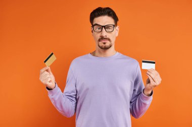 Gözlüklü ve mor kazaklı şüpheli adam turuncu arka planda iki kredi kartını karşılaştırıyor.