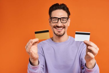 cheerful bearded man in glasses holding credit cards on orange background, shopping and consumerism clipart