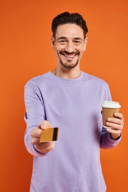 happy man in purple sweater holding coffee to go and offering credit card on orange background clipart