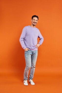 happy man in purple sweater and jeans standing with hand in pocket on orange background, casual wear clipart