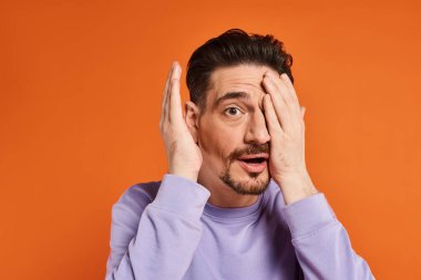 amazed man with beard in purple sweater hiding face with hand on orange background, playful mood clipart