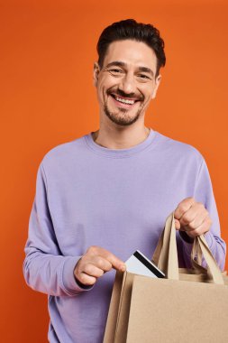 Sıradan giyinmiş neşeli sakallı adam kredi kartını turuncu arka plandaki alışveriş çantalarına koyuyor.