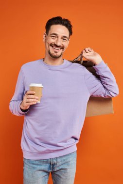 happy and bearded man holding shopping bag and coffee to go on orange background, consumerism clipart