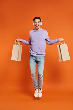 Kablosuz kulaklıklı heyecanlı adam turuncu arka planda alışveriş torbalarıyla havada uçuyor.