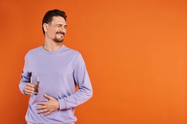Mor kazaklı sakallı adam midesi ağrıyor ve elinde turuncu arka planda bir bardak su tutuyor.