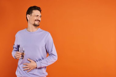 bearded man in purple sweatshirt feeling stomachache and holding glass of water on orange background clipart