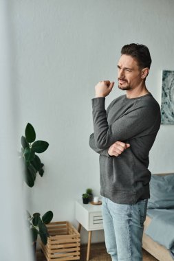 thoughtful and bearded man in grey jumper looking away while standing at home, deep in thought clipart