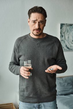 Sakallı adam elinde bir bardak su ve ilaç tutarken kameraya bakıyor.