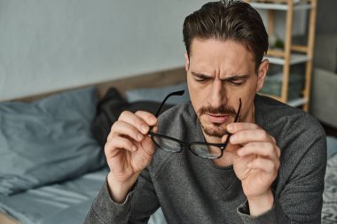 Sakallı bir adam, ciddi yüzlü ve gözlüklerine dikkatlice bakıyor.