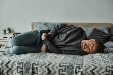 Sakallı adam yatakta yatıyor ve acıdan kaşlarını çatarken karın ağrısı, ekşitme ve acı çekiyor.