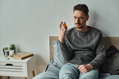 Düşünceli bir adam yatakta yatarken ilaç şişesine bakıyor. Tedavi planı.