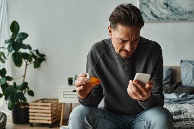 Kafası karışmış sakallı adam elinde haplarla elinde şişeyle akıllı telefonuna reçete okuyor.