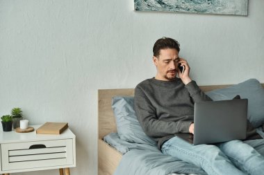 Sakallı adam bilgisayarını kullanıyor ve evden uzakta çalışırken telefonla konuşuyor, serbest çalışıyor.