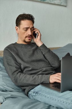 Sakallı adam bilgisayarını kullanıyor ve evden uzakta çalışırken cep telefonuyla konuşuyor.