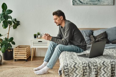Evde bilgisayarın yanında çalışırken cep telefonu kullanan sakallı bir adam.