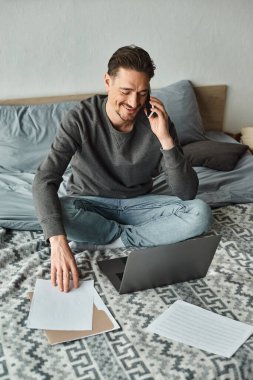 Mutlu ve sakallı bir adam evde, yatak odasında çalışırken bilgisayarını ve akıllı telefonunu kullanıyor.