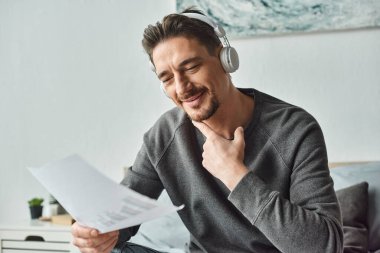 Kablosuz kulaklıklı mutlu adam, grafikleri analiz ediyor ve yatak odasında müzik dinliyor, evden çalışıyor.