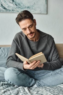 Gri kazak giyen sakallı adam hafta sonu yatak odasında dinlenirken kitap okuyor.