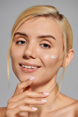 Yanağına krema sürmüş güzel genç bir kadın elleriyle çeneye dokunuyor ve gri arka planda nazikçe gülümsüyor.