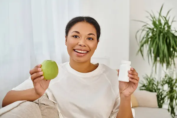 stock image happy african american dietitian holding fresh apple and supplements while smiling in living room