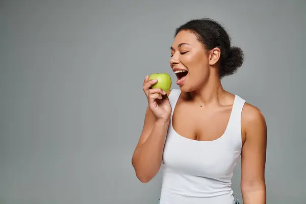 Stock image happy african american woman with white teeth biting green apple on grey background, healthy eating