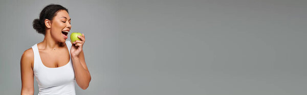 счастливая африканская американка с белыми зубами кусающая зеленое яблоко на сером фоне, баннер