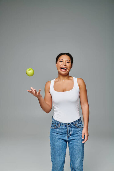 счастливая африканская американка с белыми зубами бросает зеленое яблоко в воздух на сером фоне