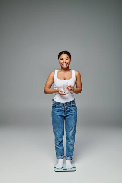 happy african american woman in casual attire holding glass of water and standing on scales on grey