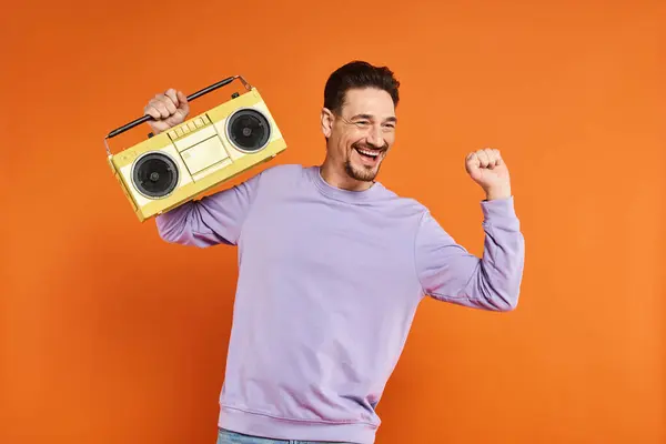 stock image joyful and bearded man in purple sweatshirt holding retro boombox on orange background, music