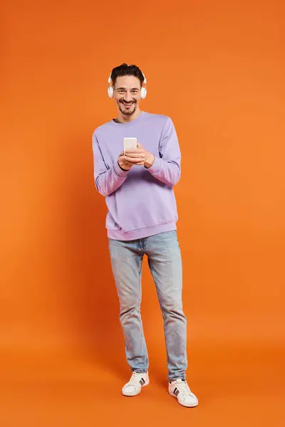 happy man in wireless headphones listening music and using smartphone on orange background