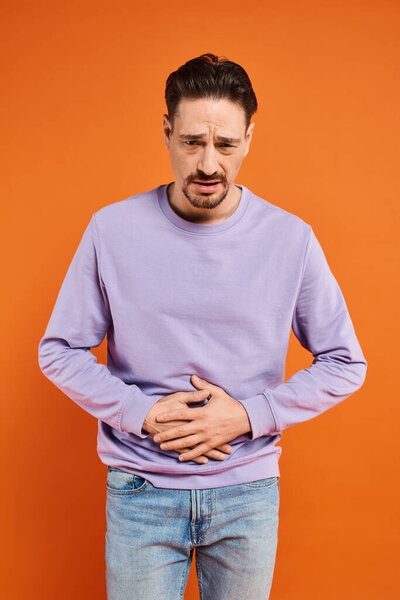 бородатый мужчина в фиолетовом свитере и джинсах, страдающих от боли в животе на оранжевом фоне, боли