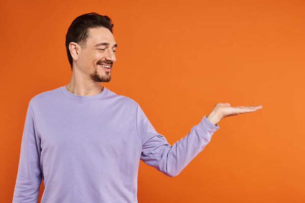 счастливый бородатый мужчина в фиолетовой толстовке представляет, указывая рукой на оранжевый фон