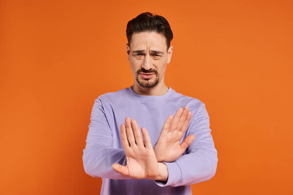 бородатый мужчина в фиолетовом свитере показывает жест отказа руками на оранжевом фоне