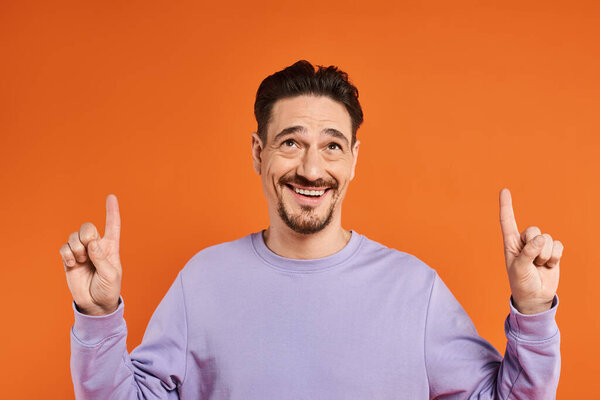 веселый мужчина в фиолетовой толстовке указывает пальцами на оранжевый фон, представляя
