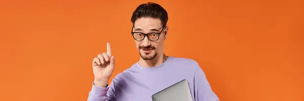 stock image bearded man in glasses and purple sweater holding laptop and pointing up on orange backdrop, banner