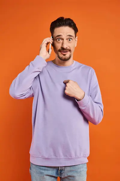 stock image surprised man talking on smartphone and pointing at himself on orange background, digital life