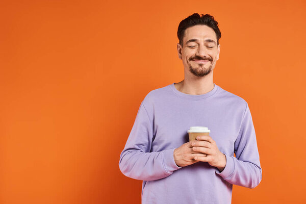 pleased and bearded man holding paper cup with coffee to go on orange background, enjoyment