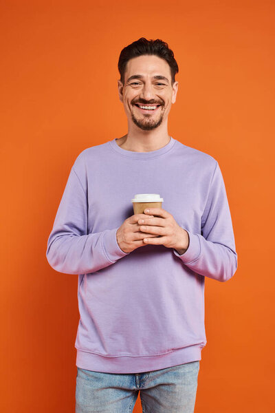 cheerful and bearded man holding paper cup with coffee to go on orange background, enjoyment