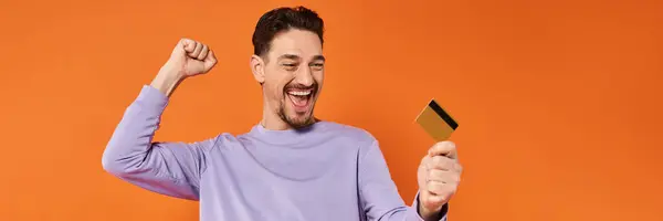 stock image excited man with beard smiling and holding credit card on orange background, rejoicing banner